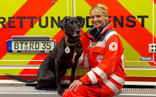 Maintrailer-Rettungshund Toto mit seiner Hundeführerin Caroline vor einem Rettungswagen.