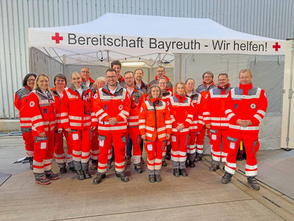 Die Kameradinnen und Kameraden der BRK-Bereitschaft Bayreuth I, die 470 Stunden lang ehrenamtlich den Festbetrieb des Maisel`s Weißbierfest in Bayreuth 2025 absicherten, Im Hintergrund die Unfallhilfsstelle auf dem Gelände des Maisel`s Weißbierfestes - ein Pavillion mit der Aufschrift "Bereitschaft Bayreuth - Wir Helfen!"