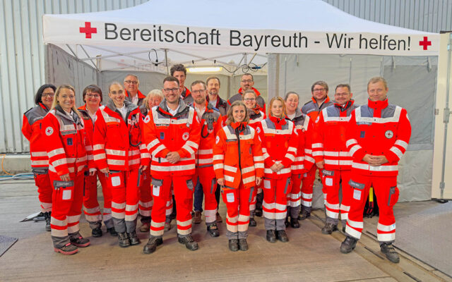 Die Kameradinnen und Kameraden der BRK-Bereitschaft Bayreuth I, die 470 Stunden lang ehrenamtlich den Festbetrieb des Maisel`s Weißbierfest in Bayreuth 2025 absicherten, Im Hintergrund die Unfallhilfsstelle auf dem Gelände des Maisel`s Weißbierfestes - ein Pavillion mit der Aufschrift "Bereitschaft Bayreuth - Wir Helfen!"