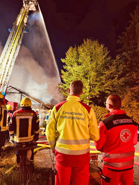 Kräfte von BRK und Feuerwehr in Bereitstellung, im Hintergrund die laufende Brandbekämpfung mit einer der eingesetzten Drehleitern.