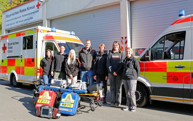 Markus, Nele, Julian und Mea starten ihre Ausbildung zum/zur Notfallsanitäter/-in beim BRK-Rettungsdienst in Bayreuth. Im Bild zusehen: Die vier neuen Azubis mit ihren Praxisanleiterinnen und -anleitern vor Fahrzeugen des Rettungsdienstes. Im Vordergrund notfallmedizinisches Equipment.