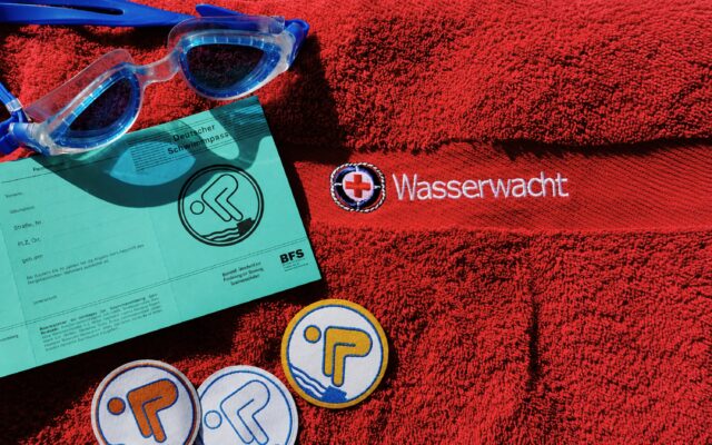 Das Bild zeigt eine rotes Handtuch mit Logo der Wasserwacht, sowie mehrere Deutsches Schwimmabzeichen, eine Schwimmbrille und einen Deutschen Schwimmpass.